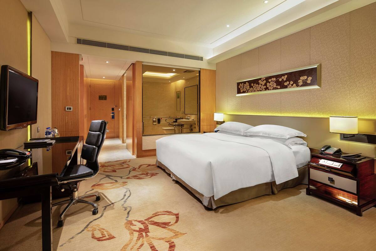 A hotel room with a bed, desk, chair, TV, bedside table, lamp, and a mirror.