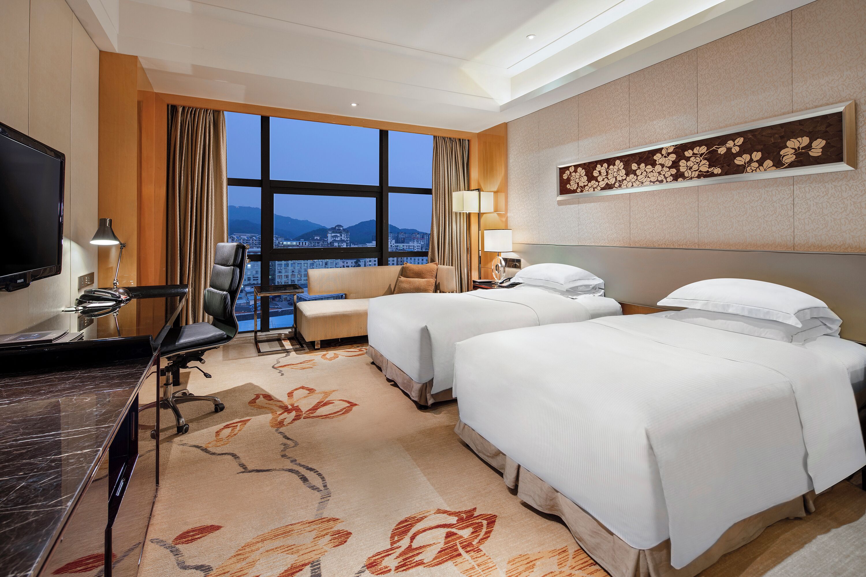 A hotel room with two beds, a desk, a TV, and a city view.