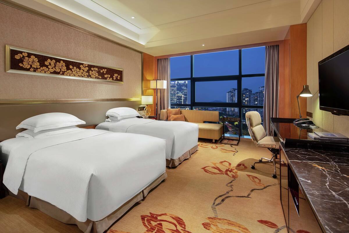 A hotel room with two beds, a desk, a TV, and a large window with a city view.