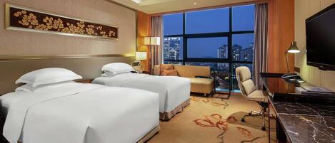 A hotel room with two beds, a desk, a TV, and a large window with a city view.