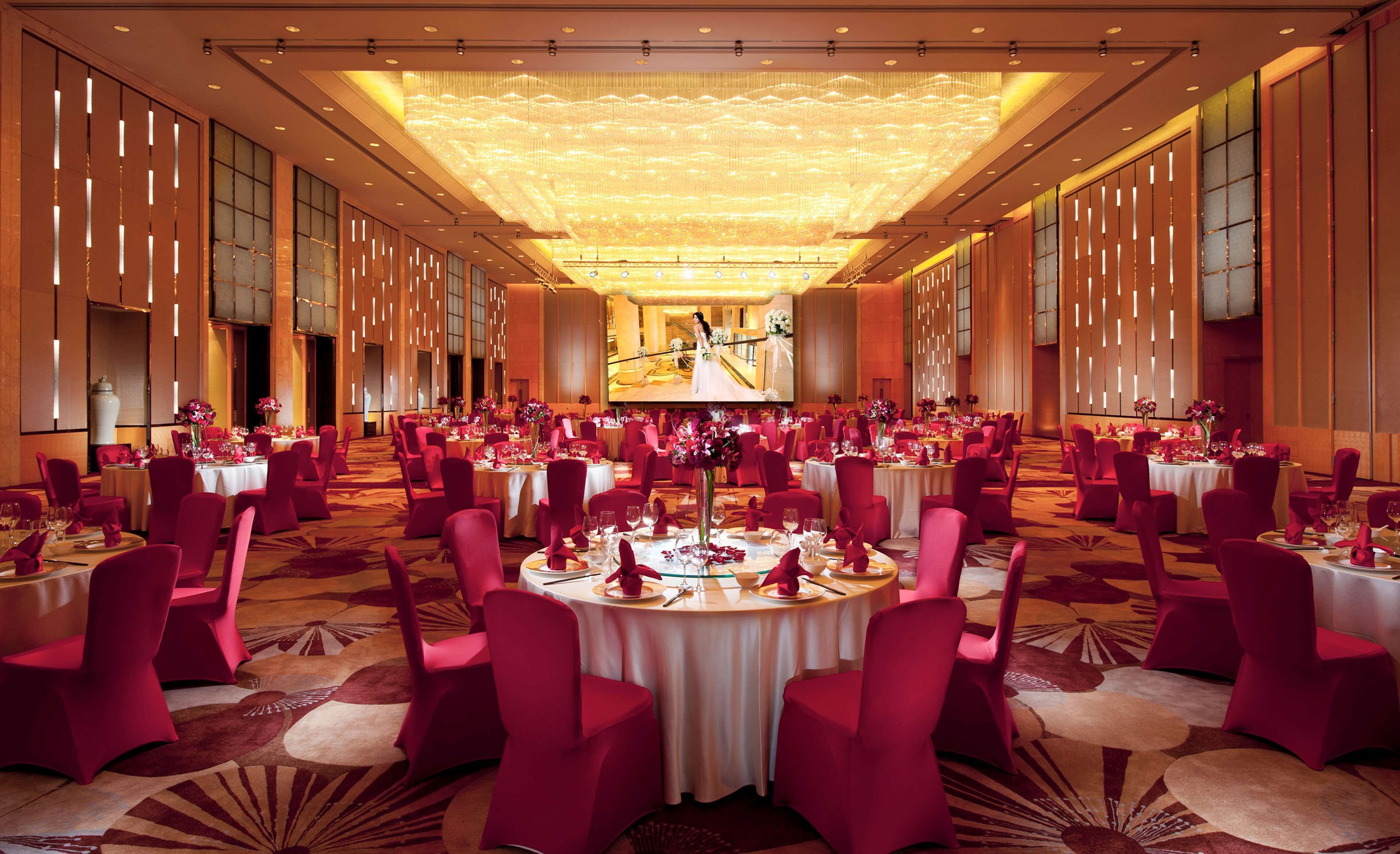 A spacious banquet hall with round tables, red chairs, and a large screen displaying a wedding scene.