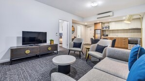 A modern living room with a flat-screen TV, a sofa, a coffee table, and a small side table. There is a kitchen area with cabinets and a microwave.