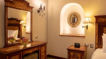 A hotel room with a wooden vanity, a large mirror, a bedside table with a lamp, a decorative wall mirror, and a bed with white linens.