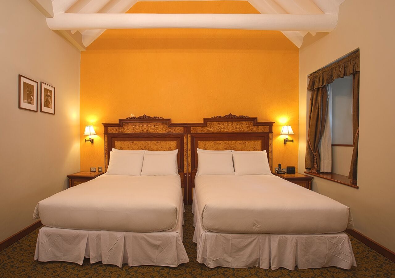 A hotel room with two beds, a wooden headboard, bedside lamps, a mirror, and a window with curtains.