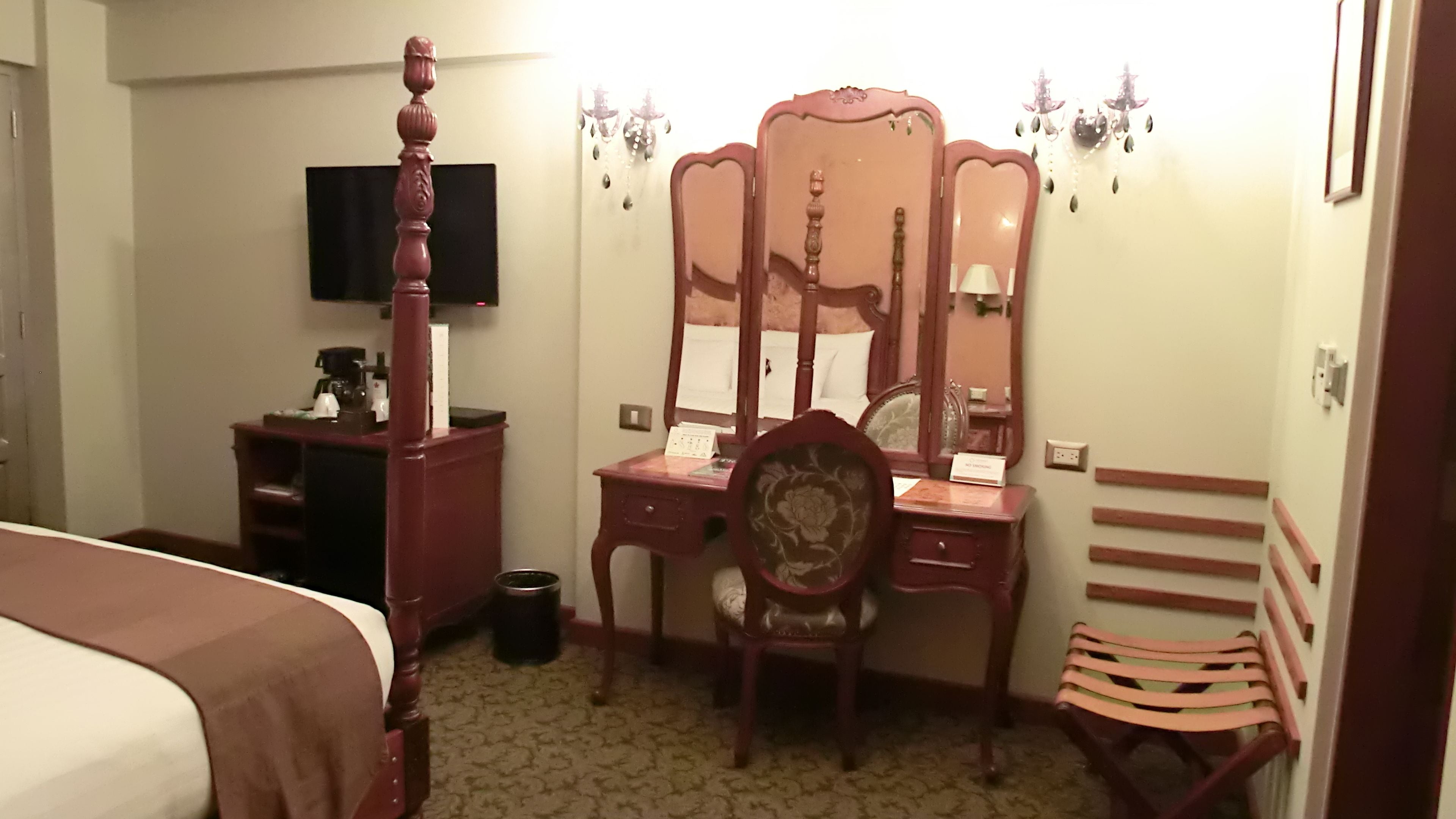 Habitación de hotel con una cama, un escritorio de madera con un espejo, una silla, una mesita con teléfono y un televisor en la pared.