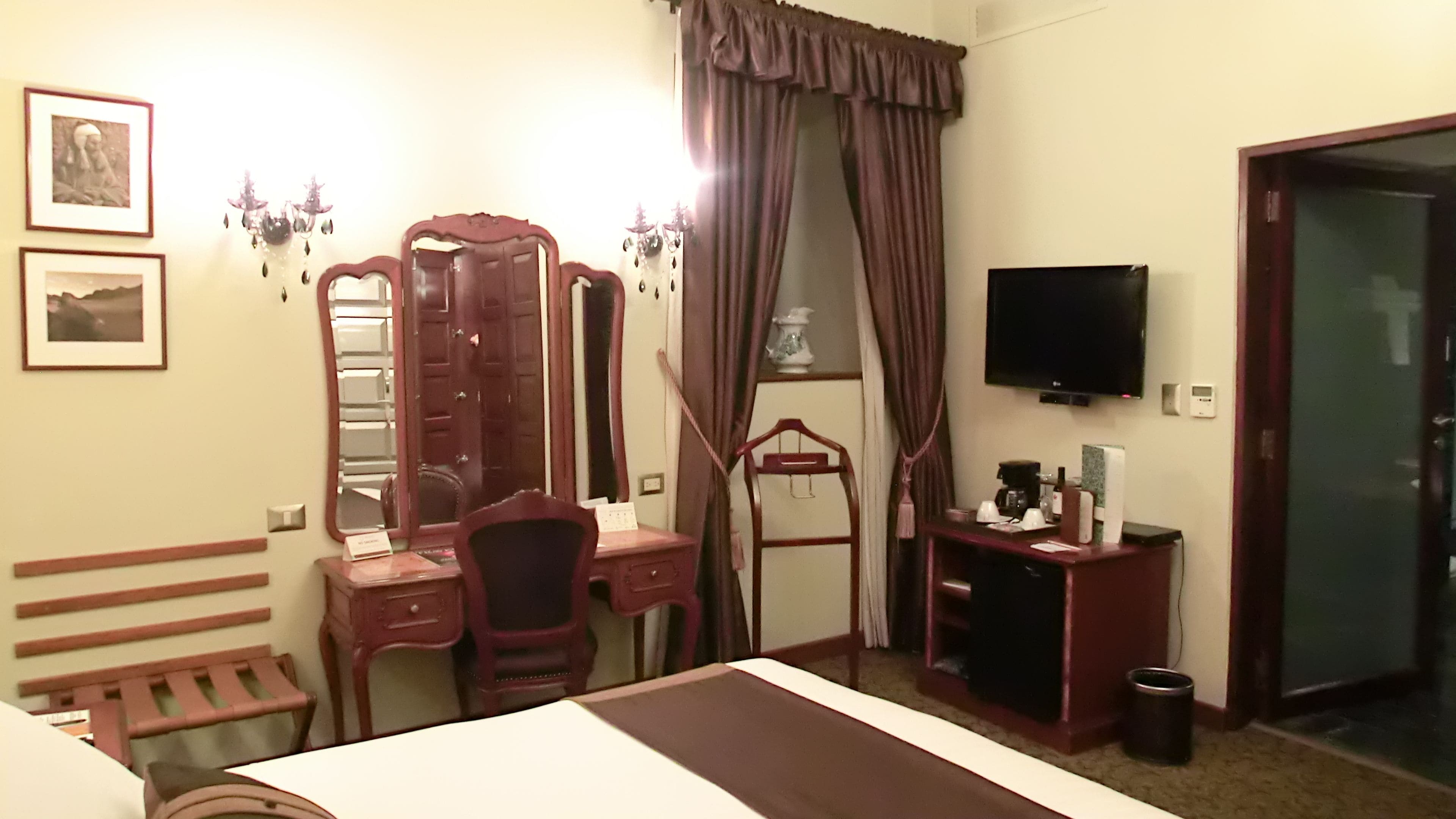 A hotel room with a bed, a desk, a chair, a vanity with a mirror, a television, and a door.
