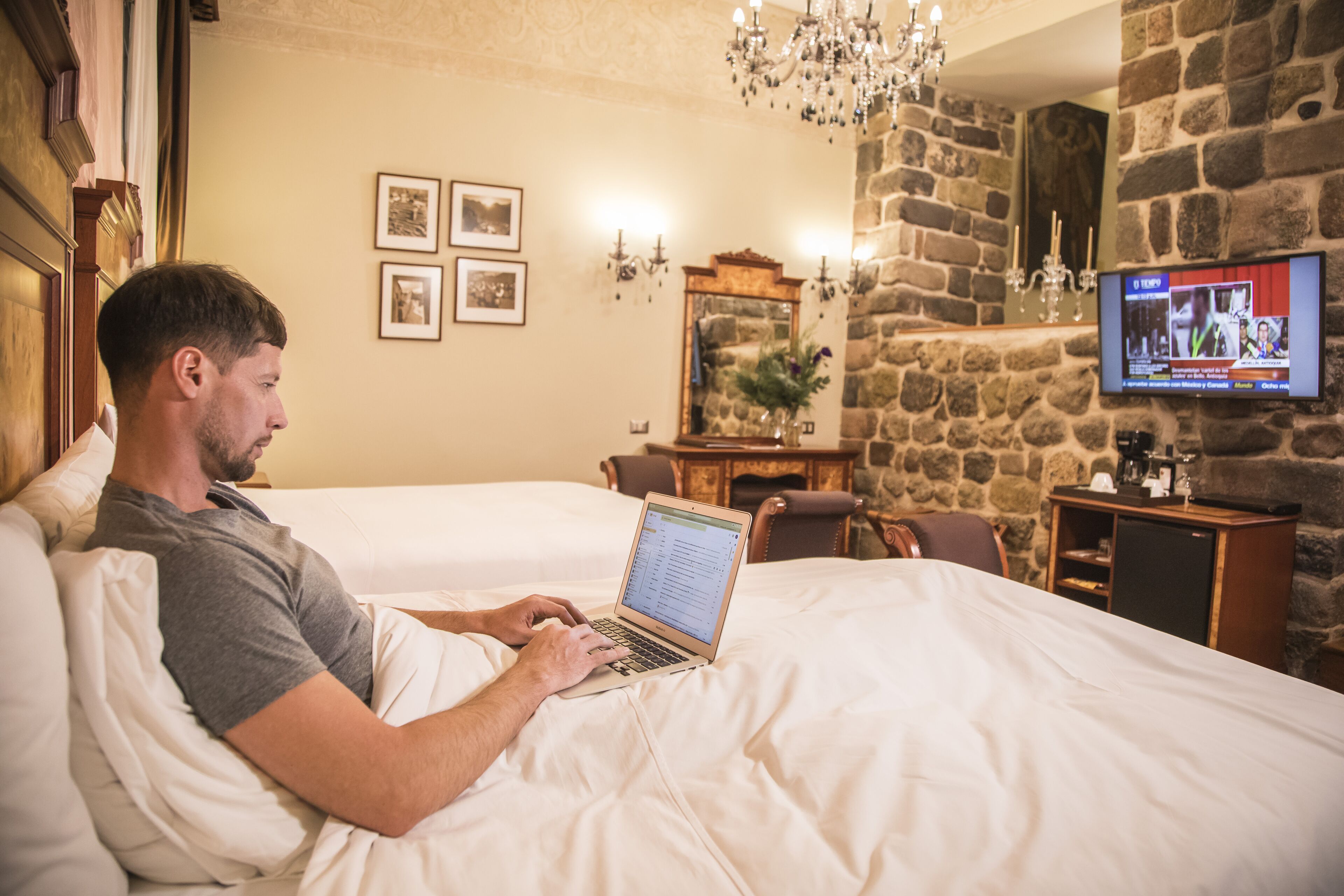 Un hombre usando una computadora portátil en una cama dentro de una habitación de hotel con pared de piedra, un televisor y cuadros enmarcados.