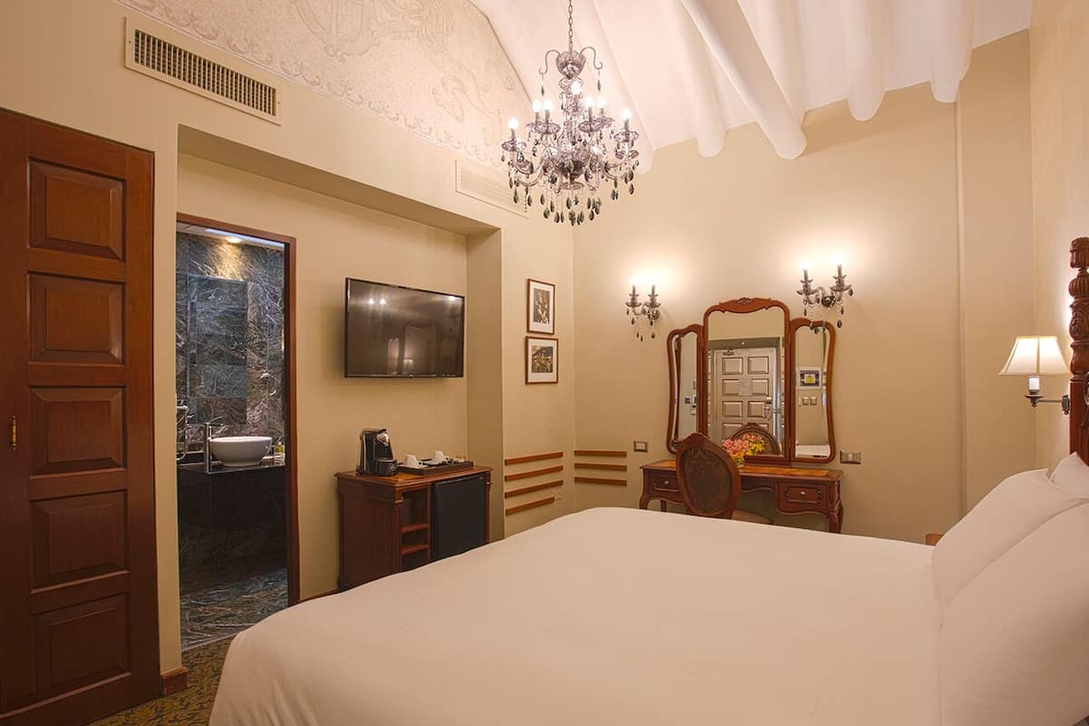 Een hotelkamer met een groot bed, een televisie, een kroonluchter, een spiegel en een houten garderobe.