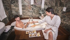 A man and a woman in a bathroom with a marble wall, a bathtub, and a tray of chocolates.