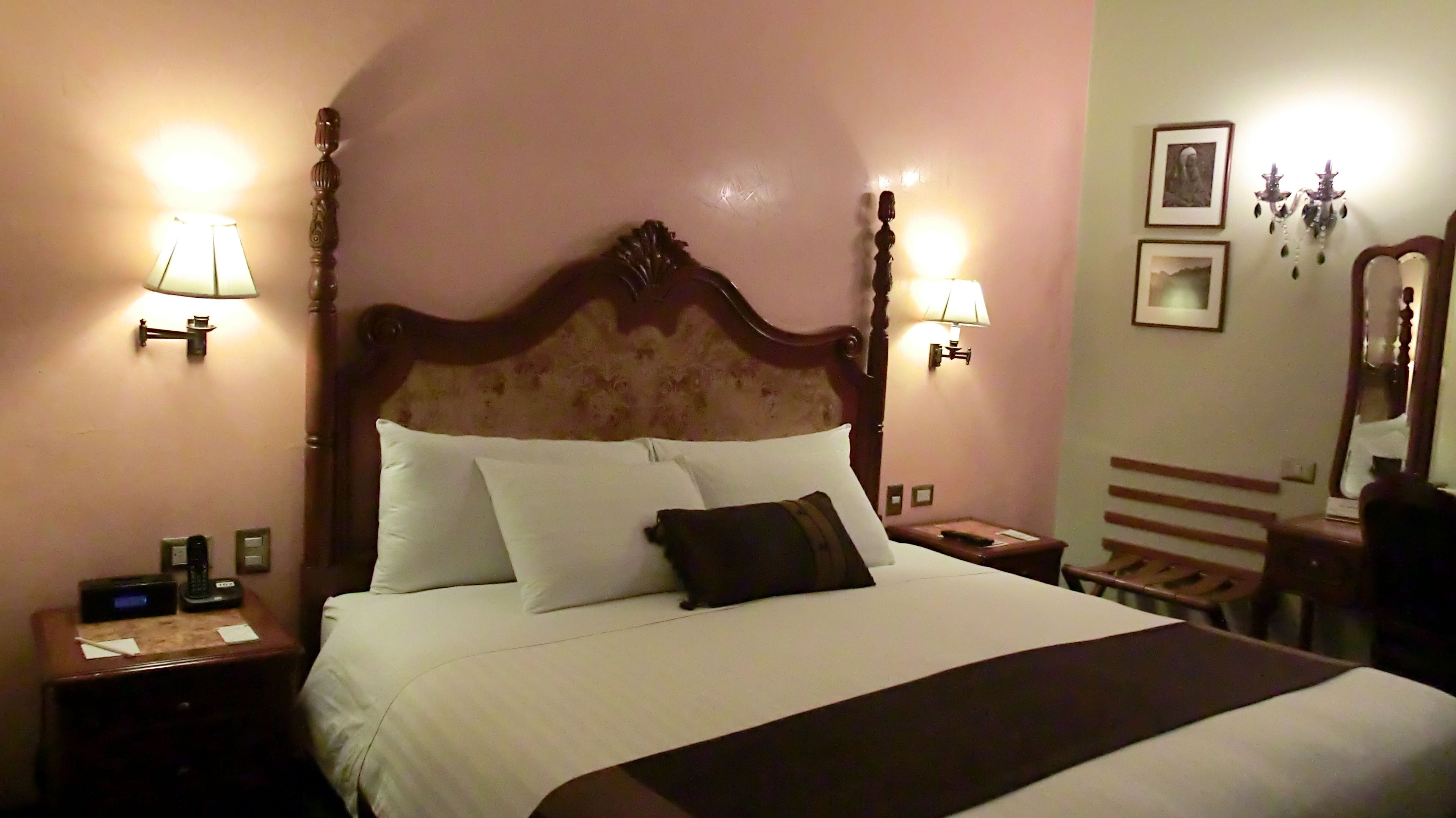A neatly made bed with white linens and a dark throw blanket. There are two bedside tables with lamps and a small clock on one. The room has a wooden headboard and a mirror on the wall.