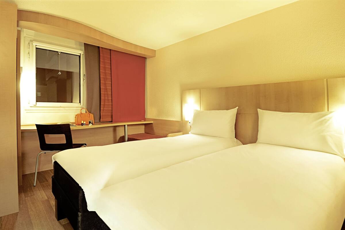 Quarto de hotel com cama de casal, uma escrivaninha com cadeira, janela com persianas e mesa de cabeceira com luminária.