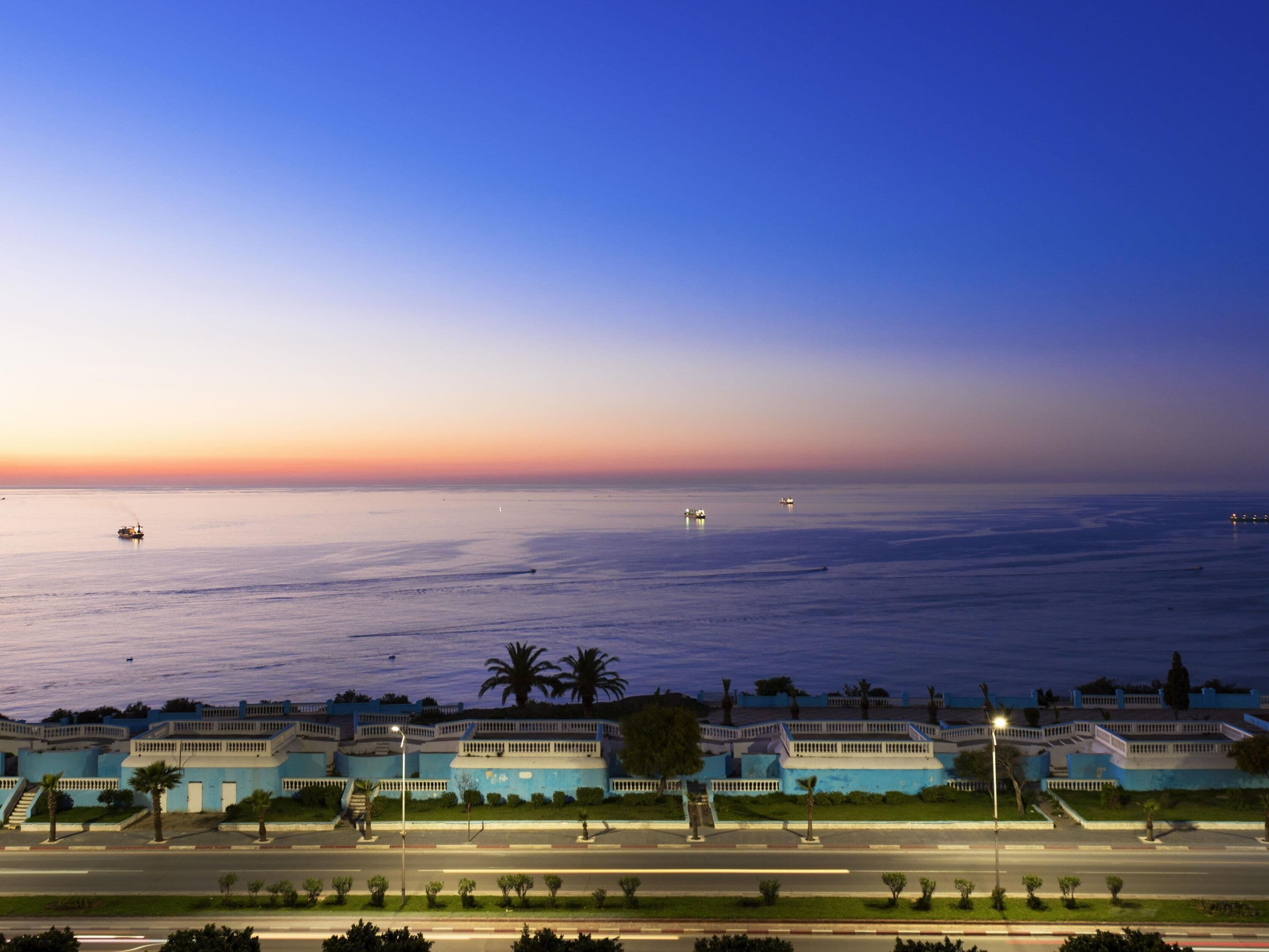 Vista costera con una carretera que lleva a varios edificios de fachadas azules y palmeras.