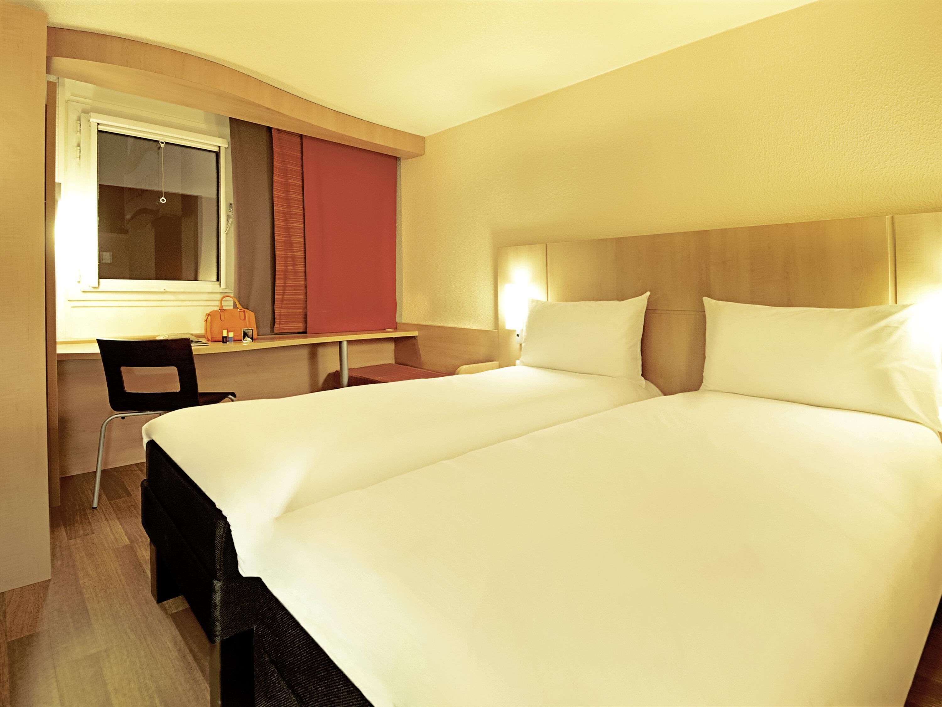 Habitación de hotel con cama doble, escritorio con silla, ventana con persianas y mesita de noche con lámpara.