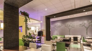 Ein modernes Restaurant mit klarem, minimalistischen Interieur, das grüne und weiße Sitzgelegenheiten, eine zentrale Bar und dekorative Bäume an der Wand zeigt.