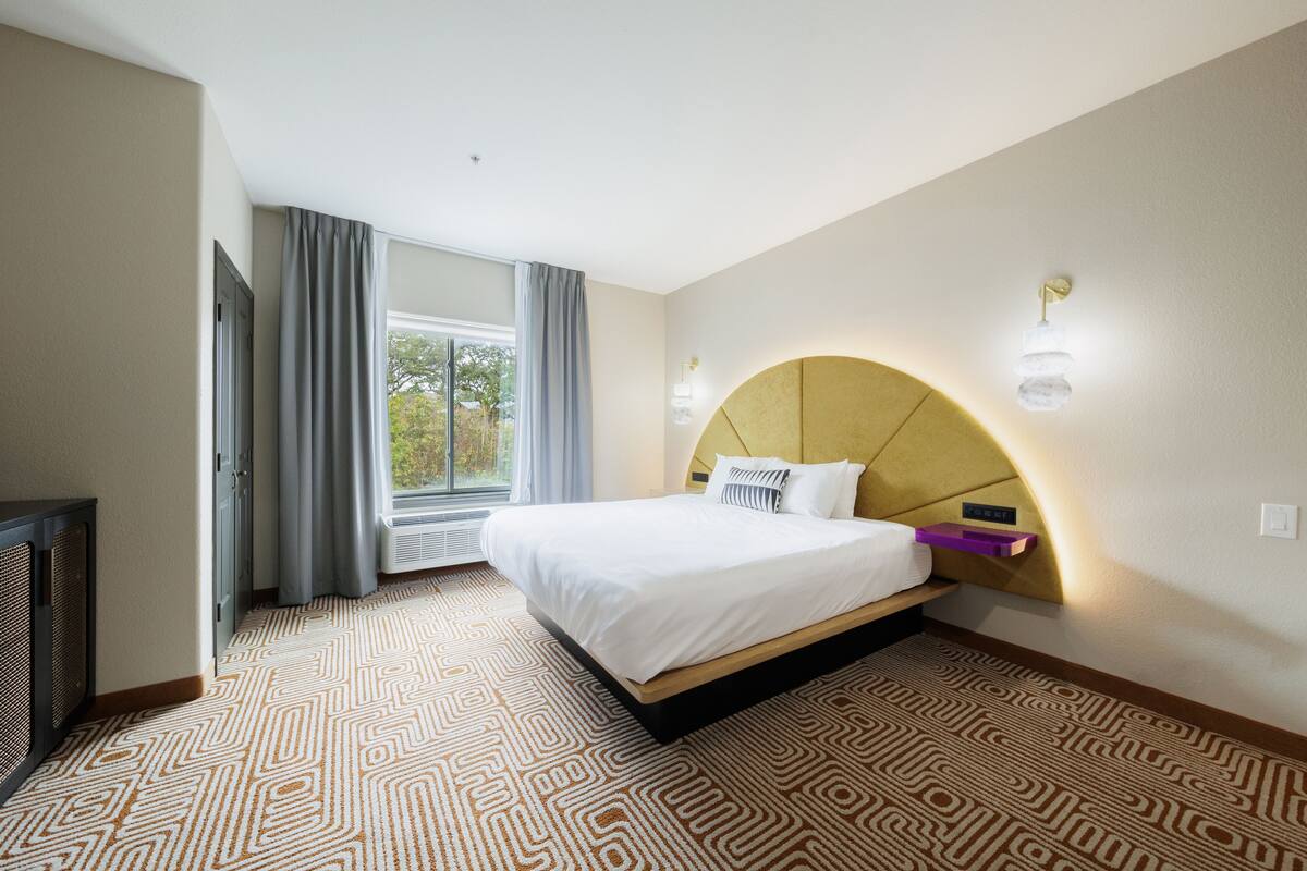 A hotel room with a large bed, a built-in TV, a window with curtains, and a patterned carpet.