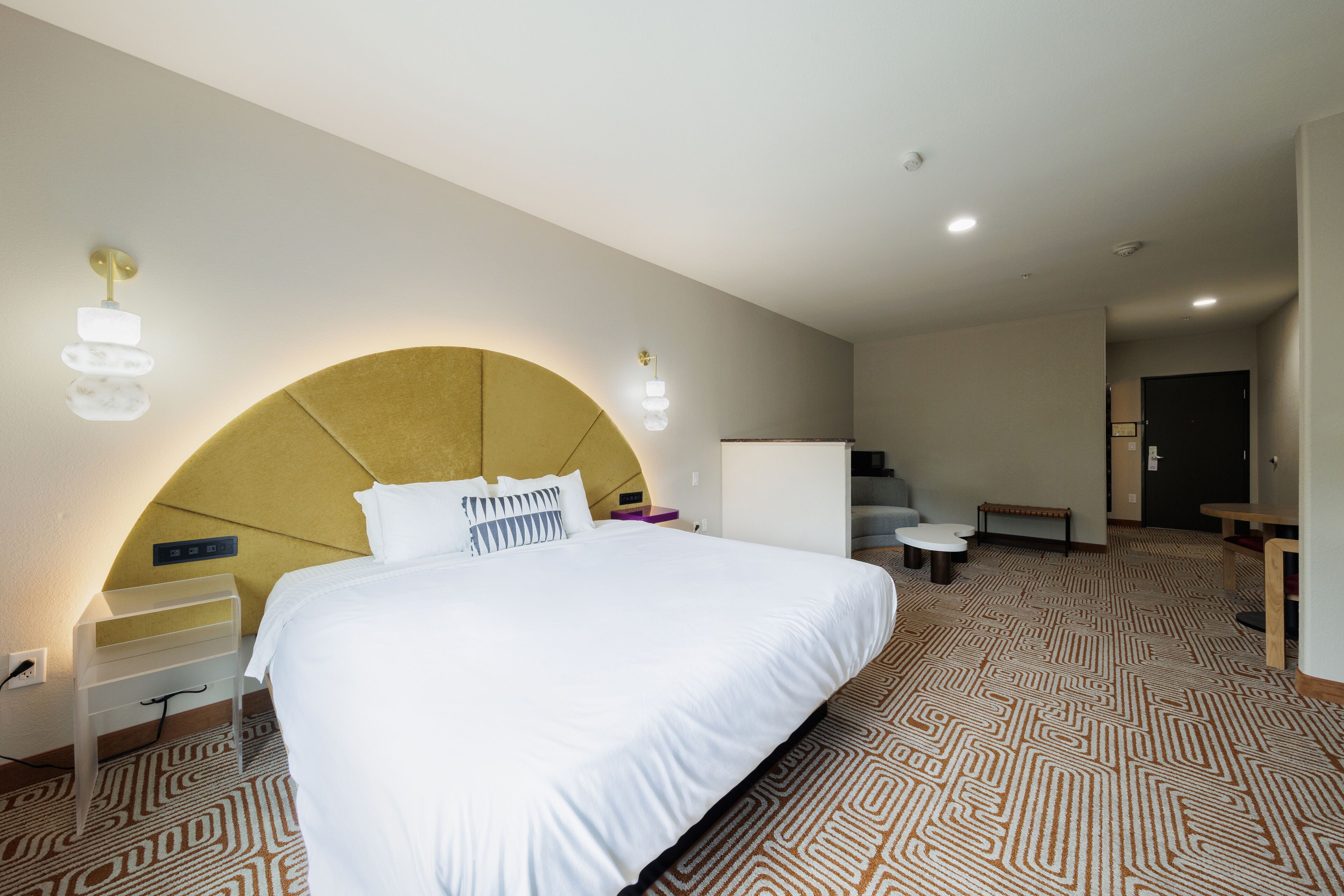 A hotel room with a large bed, a curved headboard, bedside tables, a desk, and a patterned carpet.