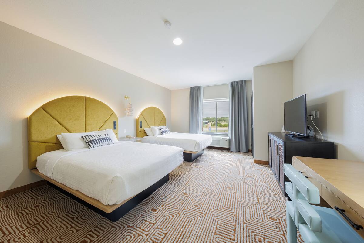 A hotel room with two beds, a desk, a television, and a large window with curtains.