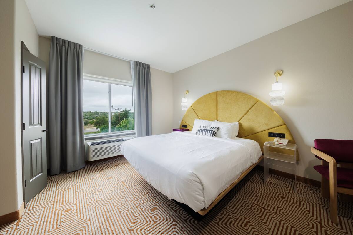 A hotel room with a large bed, a bedside table, a chair, a window with curtains, and a view of greenery outside.