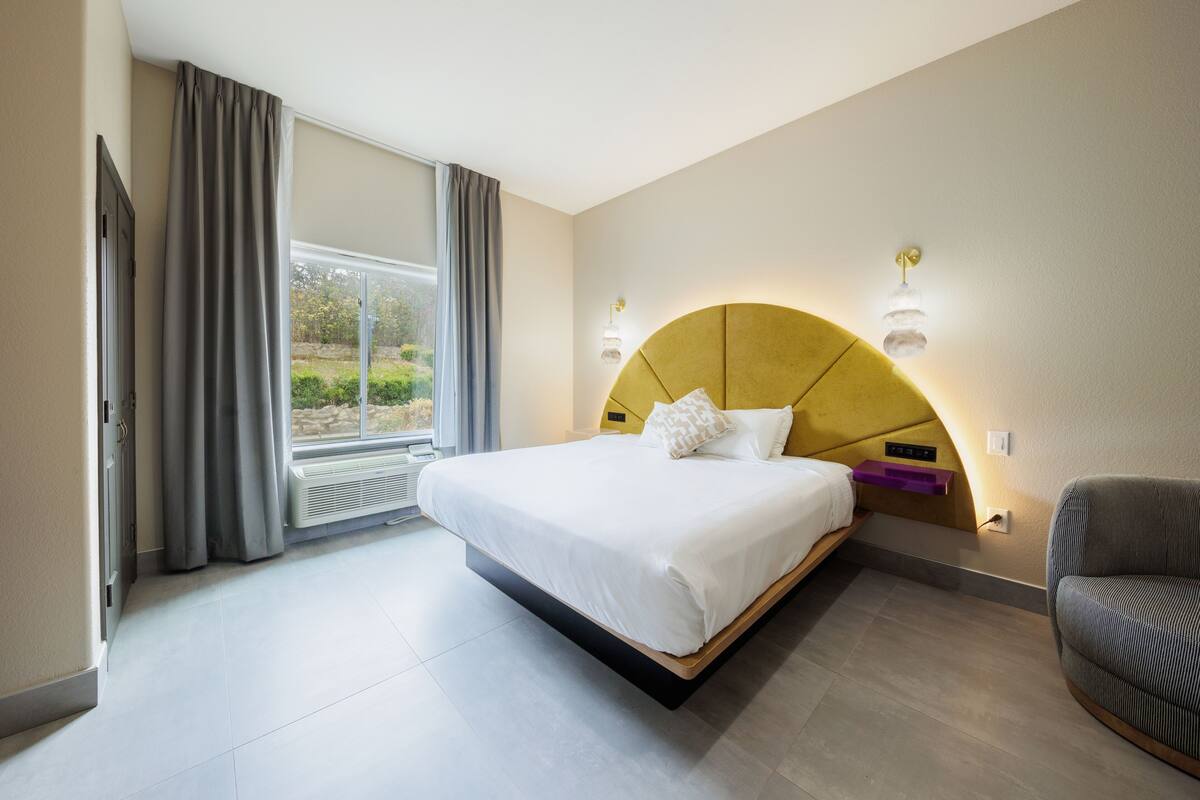 A modern hotel room with a large bed, a sofa, a window with curtains, and a built-in heater.