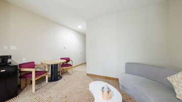 A compact living space with a sofa, a small round table, and two chairs. A compact kitchenette is visible with a microwave and a small fridge. The floor features a geometric patterned carpet.