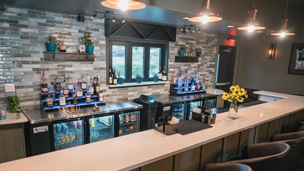 Een bar met een lange toonbank, meerdere planken vol flessen en een koelkast. Er staan potplanten op de planken en een raam met uitzicht.