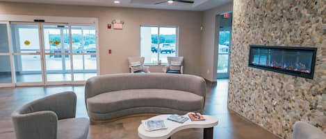 A modern lobby with a curved gray sofa, two armchairs, a coffee table, a fireplace, and a large window with a view of parked cars.