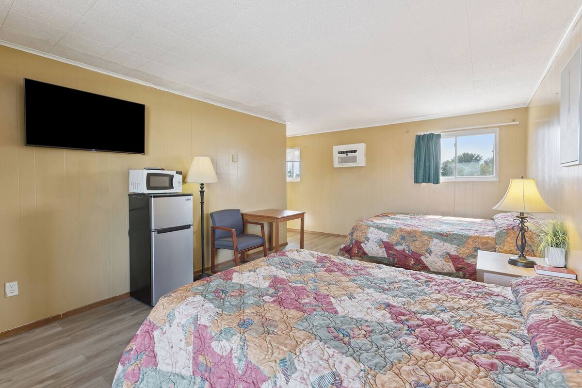 A hotel room with two beds, a desk, a microwave, a refrigerator, a TV, and an air conditioning unit.