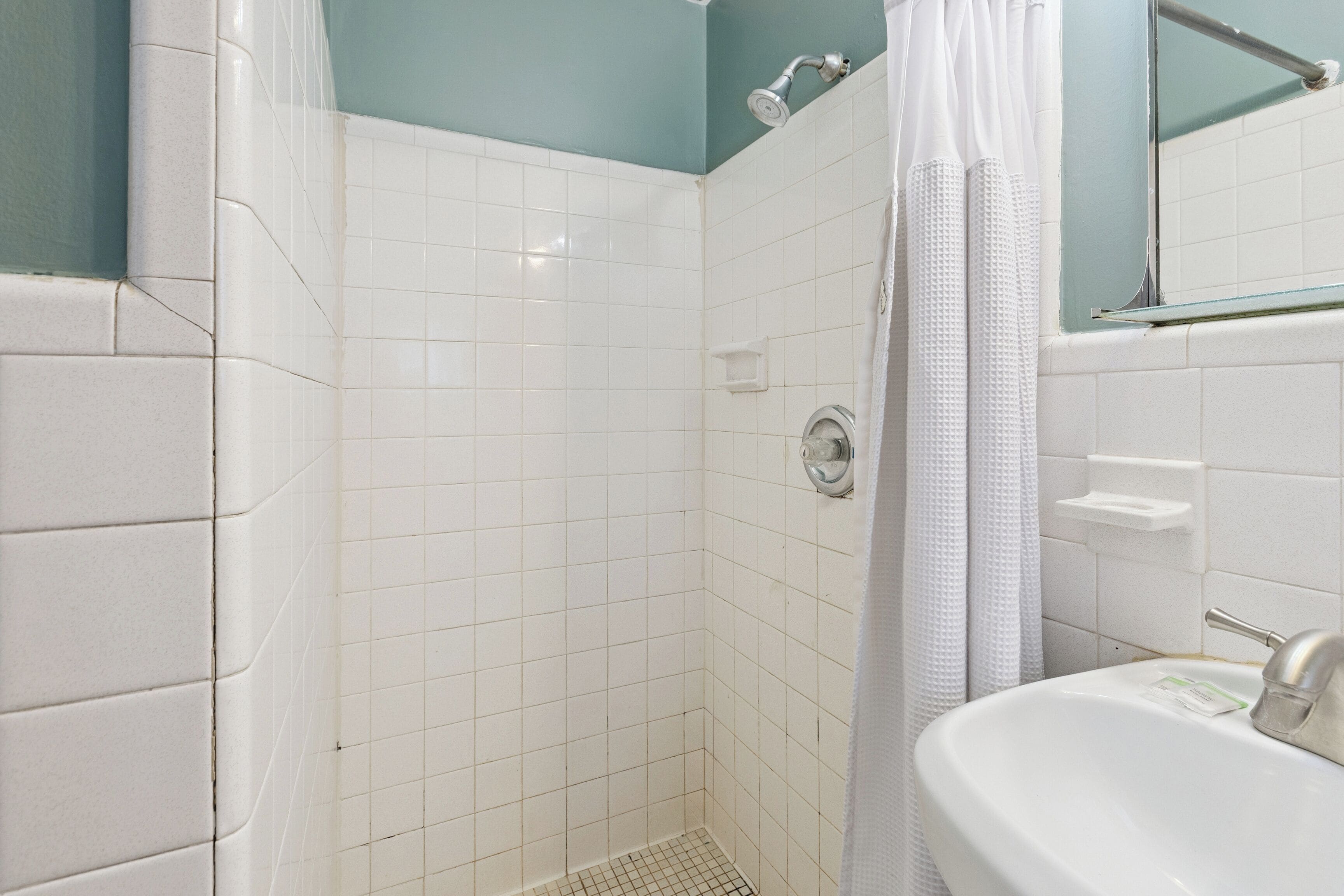 A bathroom with white tiles, a shower, a sink, and a mirror.