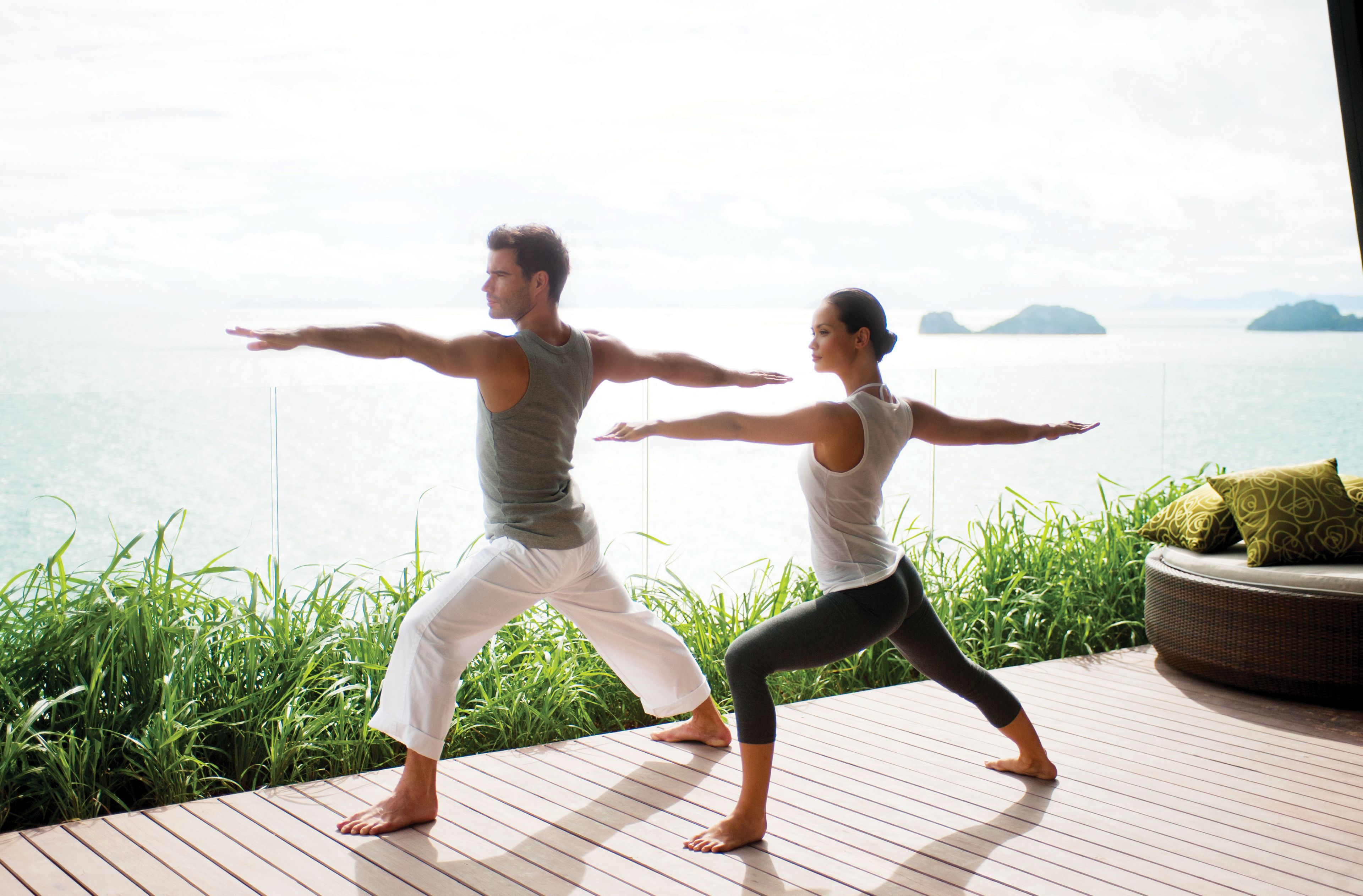 Two people practicing yoga on a wooden deck with a view of the sea and islands.
