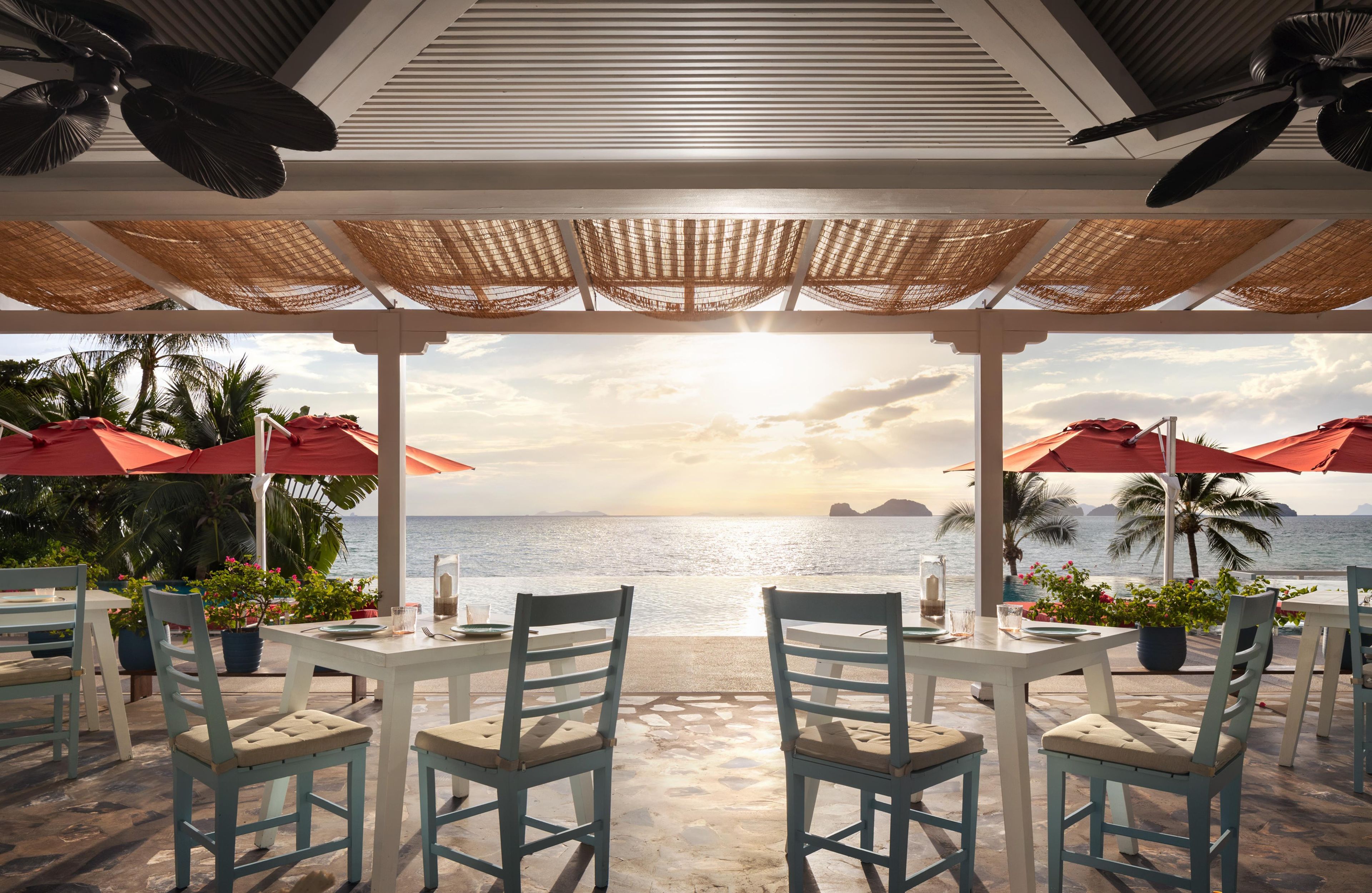 Un restaurante frente al mar con mesas y sillas, vista al océano y un atardecer.