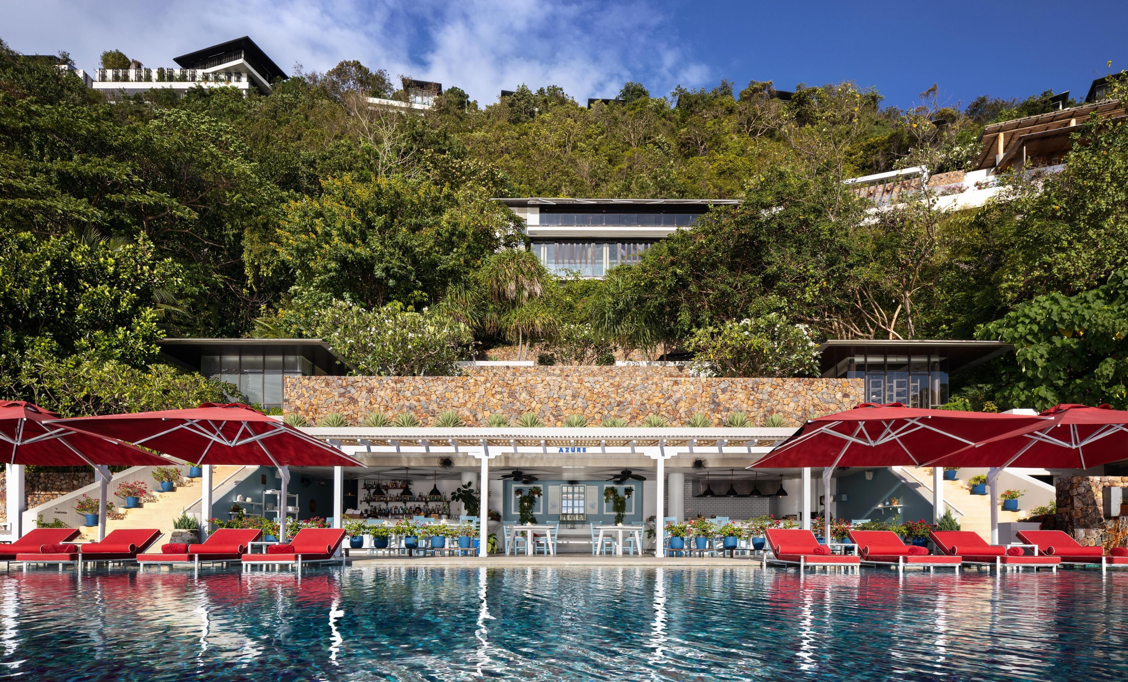 A pool area with red lounge chairs and umbrellas, a bar with a stone wall, and a view of a hillside with buildings.