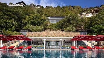 A pool area with red lounge chairs and umbrellas, a bar with a stone wall, and a view of a hillside with buildings.