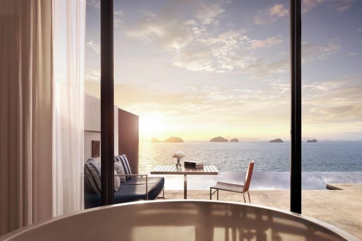 Een kamer met een groot raam met uitzicht op een strand bij zonsondergang.