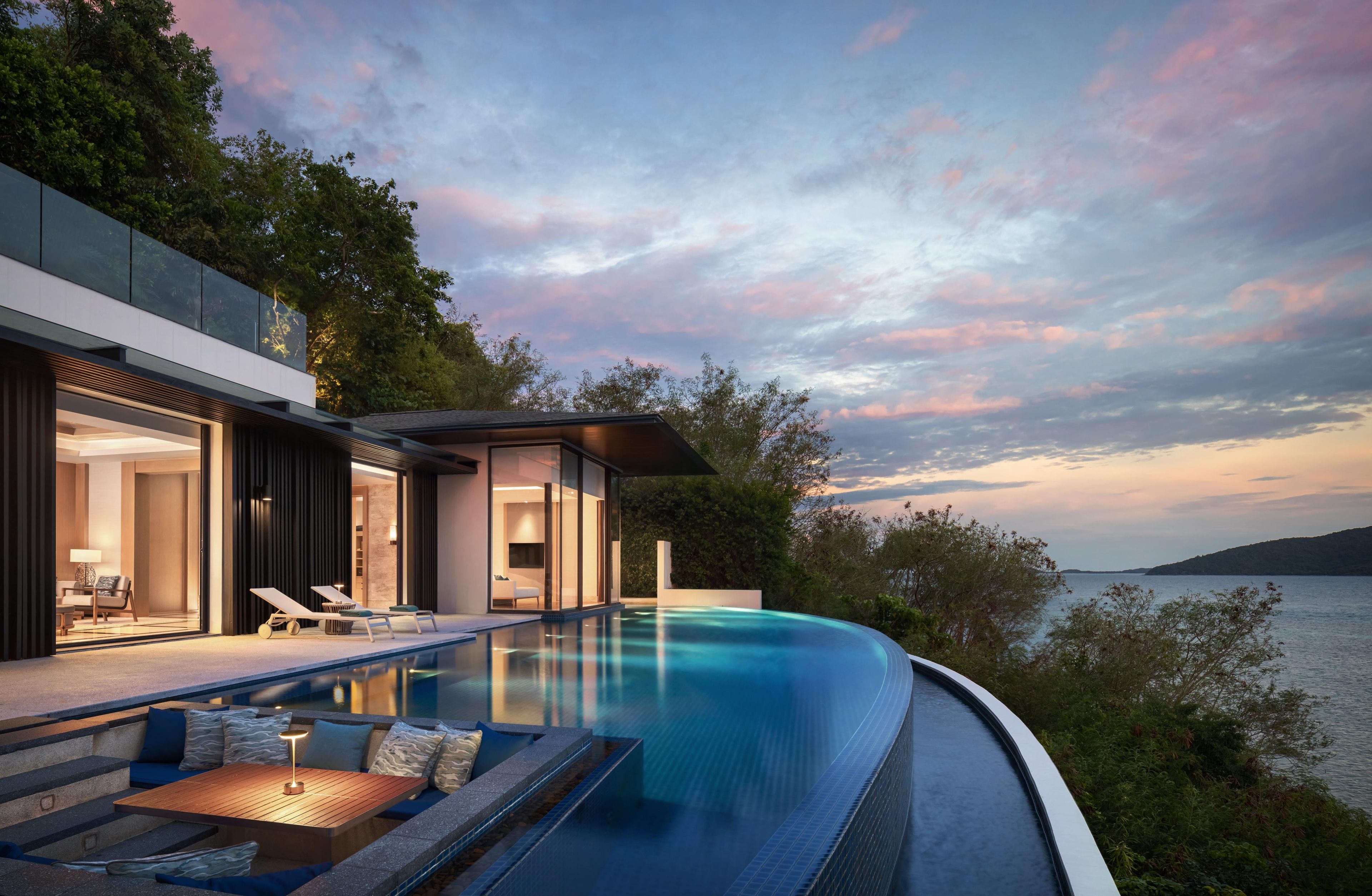 Una piscina infinita moderna con vistas al mar, un área de estar al aire libre con paredes de cristal y un edificio contemporáneo.