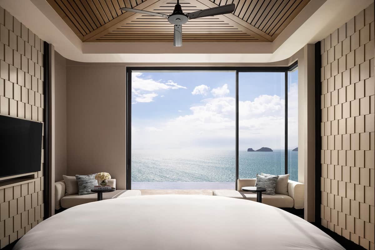 A modern hotel room with a large bed, two seating areas, a ceiling fan, and a large window offering a sea view.