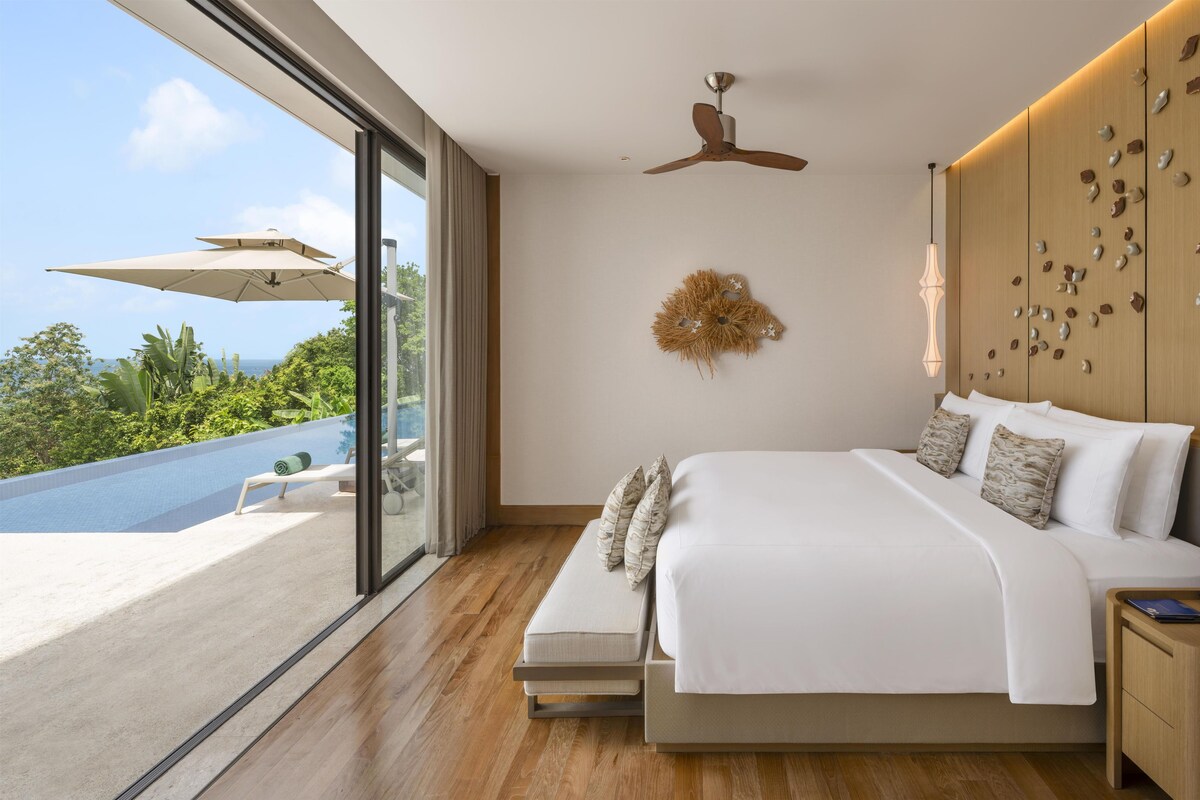 A modern bedroom with a large bed, a ceiling fan, a wall-mounted light fixture, and a view of a pool.