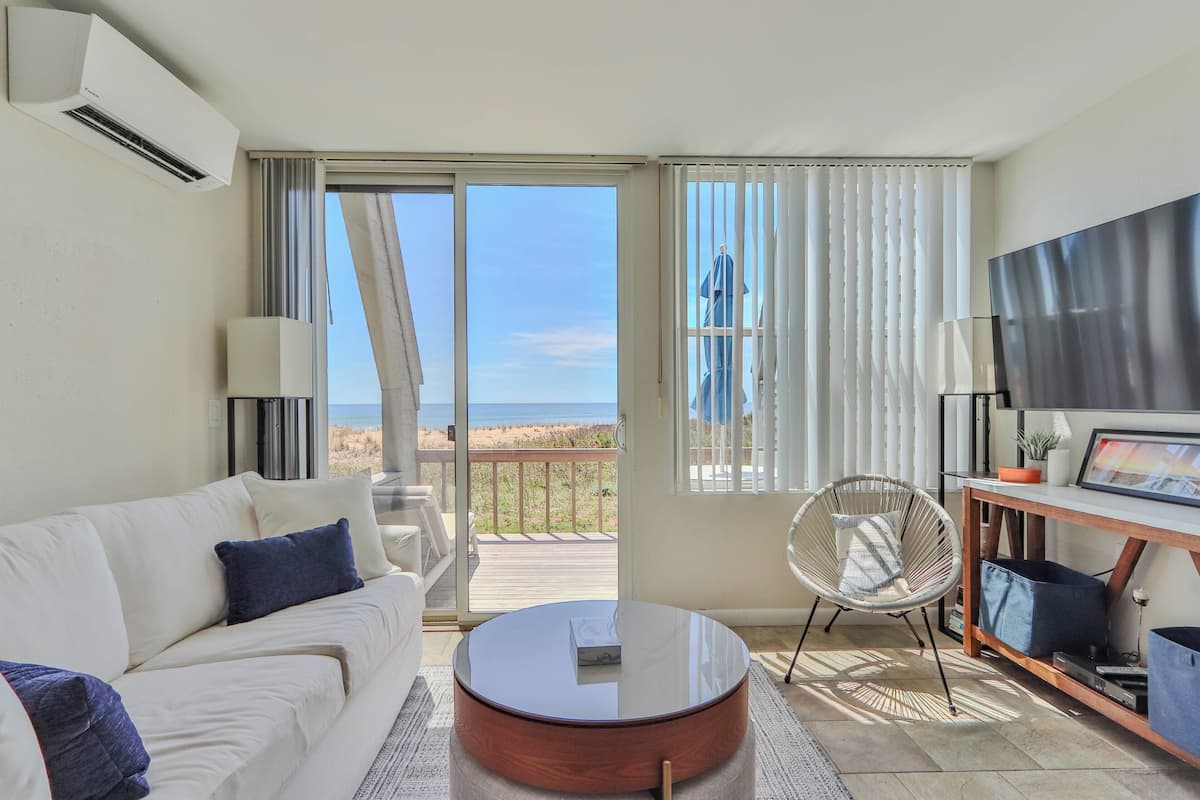 A modern living room with a white sofa, a round coffee table, a TV stand, and a view of the beach through large sliding glass doors.