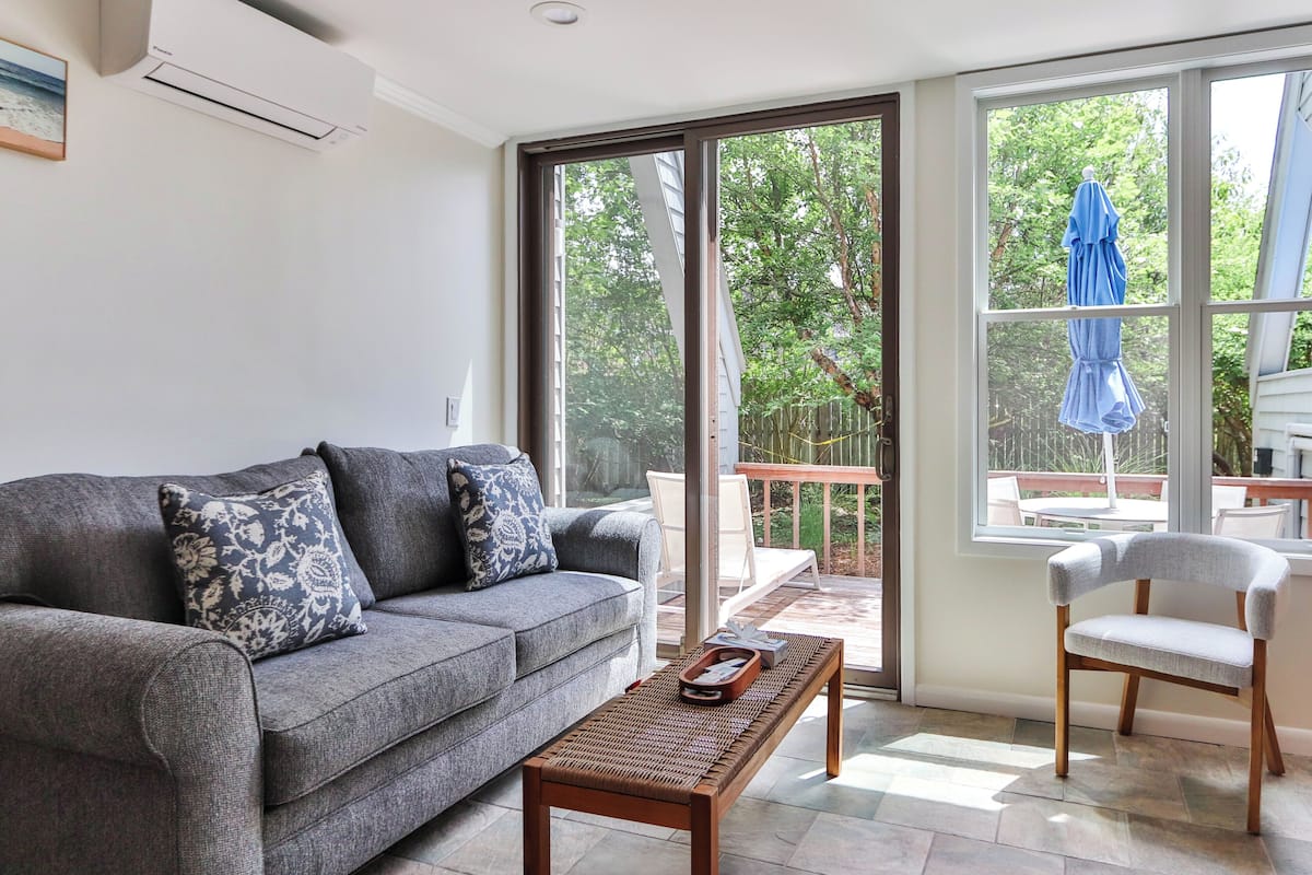 A living room with a grey sofa, a wooden coffee table, a white armchair, and a large sliding glass door leading to an outdoor patio.