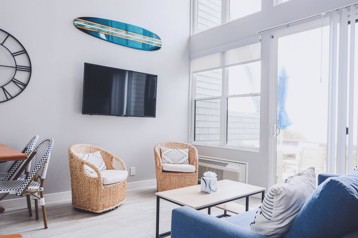 A living room with wicker chairs, a sofa, a flat-screen TV, and a large window.