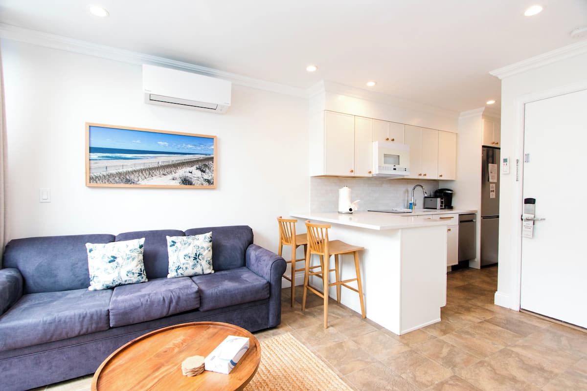 A modern living room with a sofa, a coffee table, and a kitchen area with a refrigerator, microwave, and bar stools.