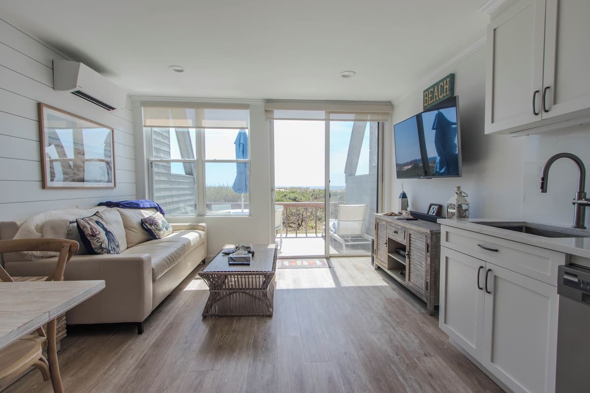 A modern kitchen with a flat-screen TV, a dining area with a table and chairs, a sofa, and a balcony with a view.
