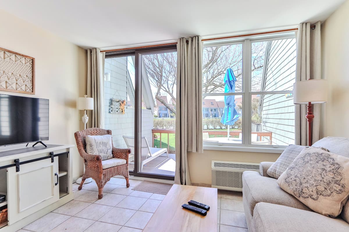 A living room with a TV, a wicker chair, a sofa, a coffee table, and a large window with a view of the outside.