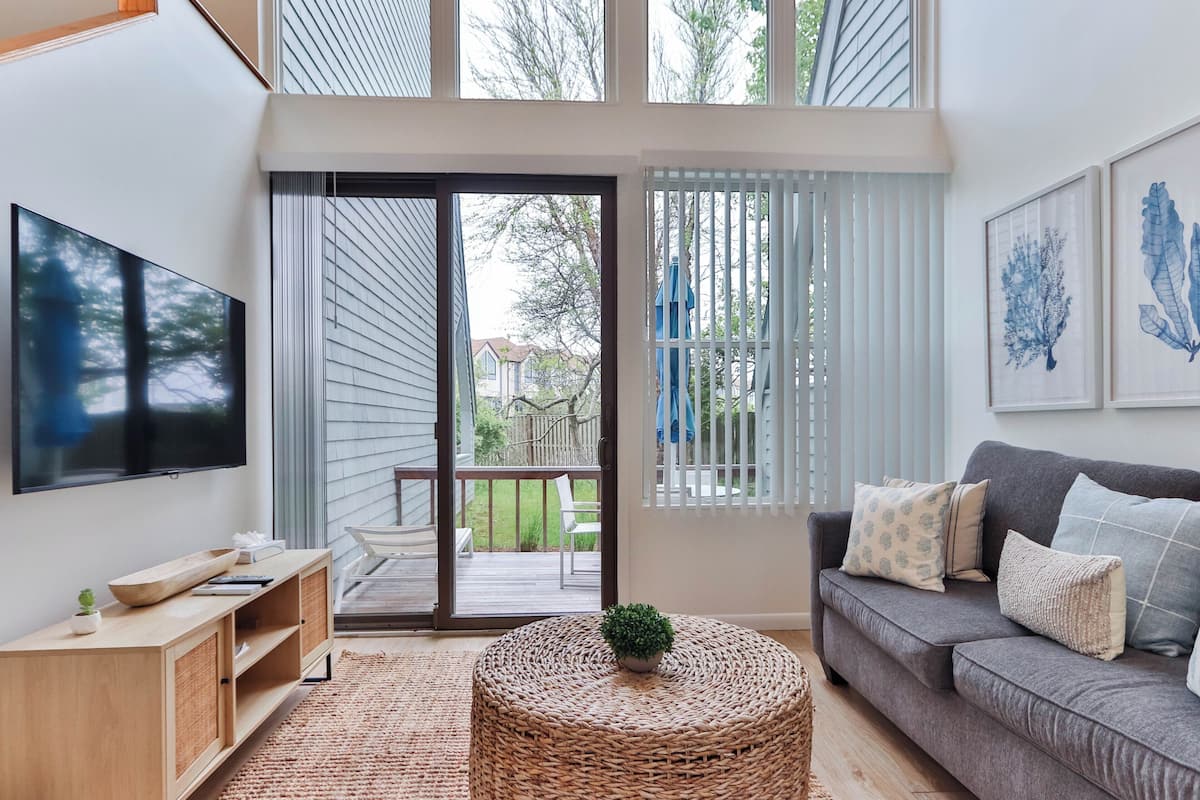 A modern living room with a flat-screen TV, a grey sofa, a wicker ottoman, and a sliding glass door leading to an outdoor deck.