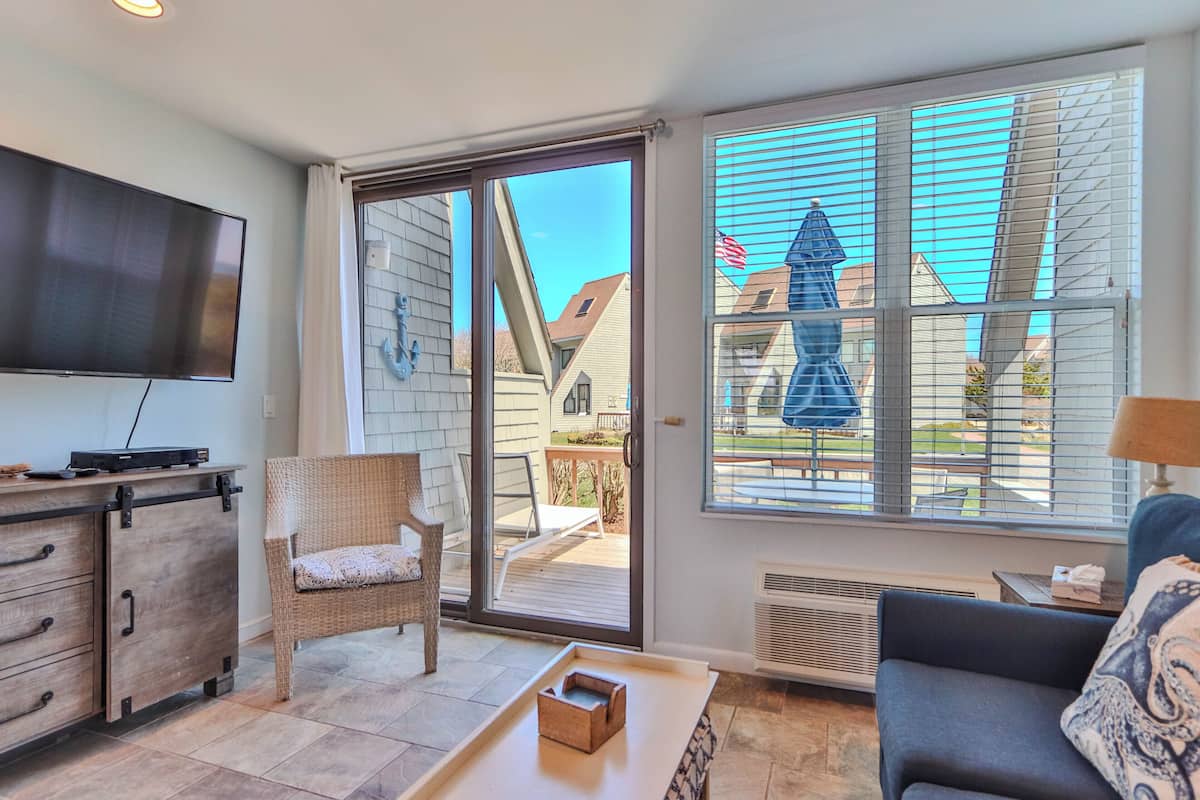 A living room with a TV, wicker chair, sofa, and a view through a sliding glass door to the outside.