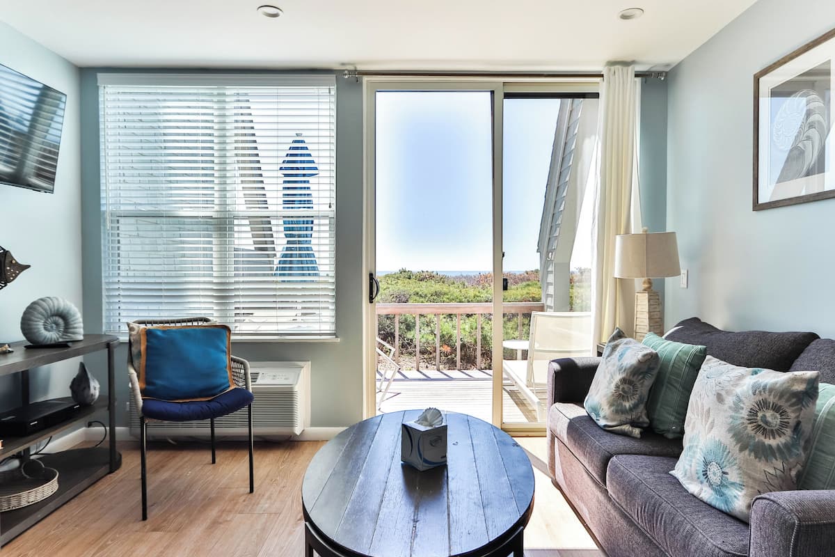 A living room with a sofa, a chair, a table, and a view through a sliding glass door to an outdoor deck.