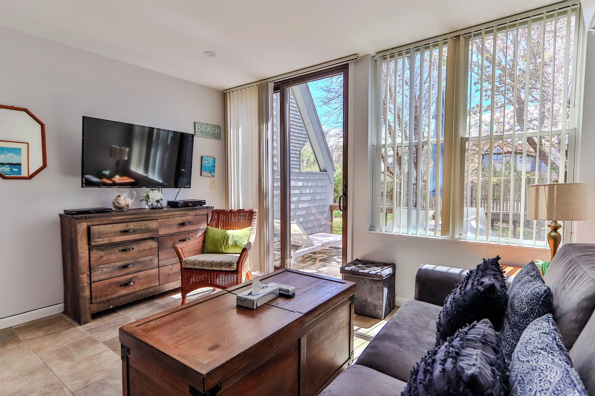 A living room with a TV, wooden furniture, a sofa, and a view through large windows.