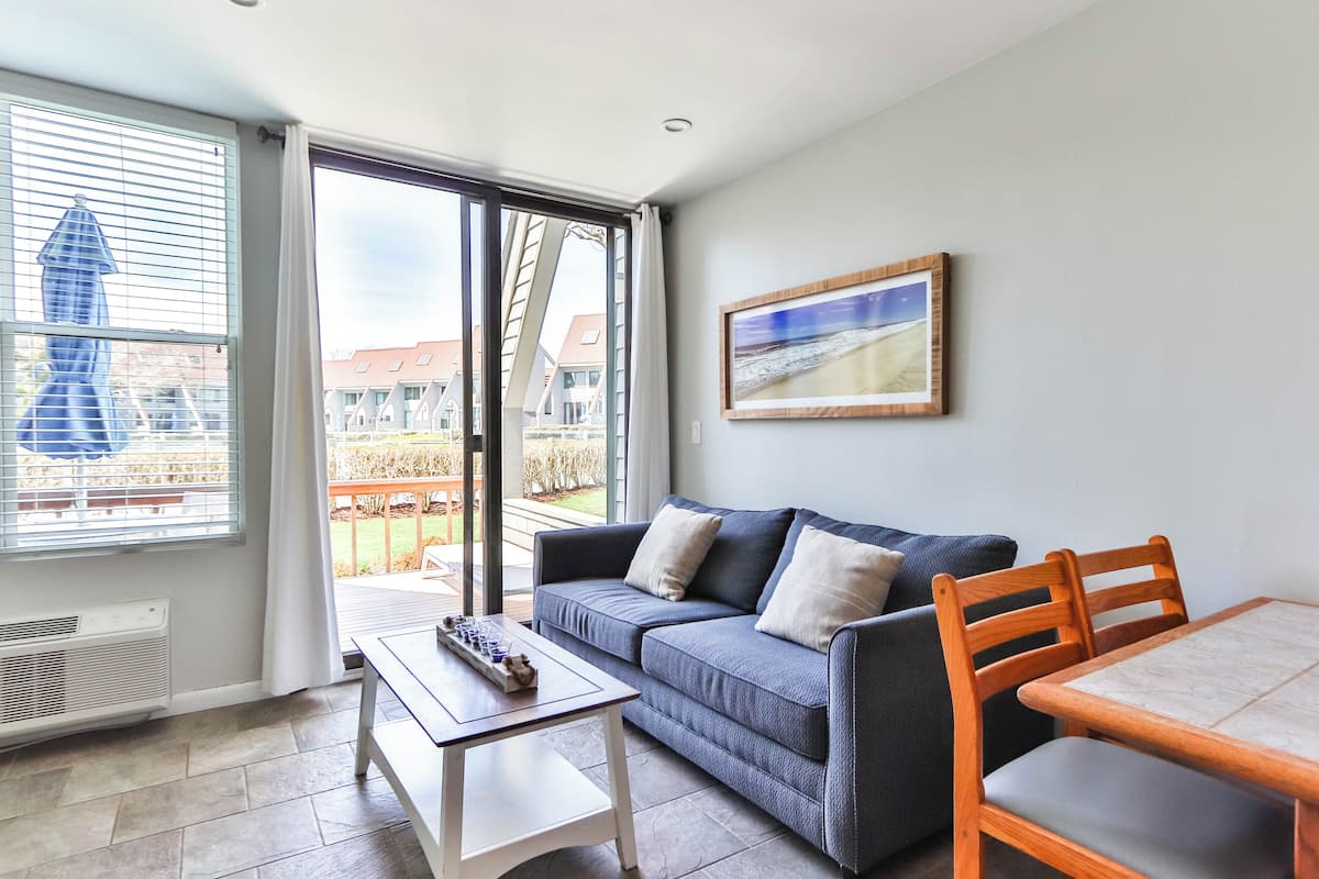 A modern living room with a sofa, a table, and a chair. There is a large window with blinds, a painting on the wall, and a view of buildings outside.