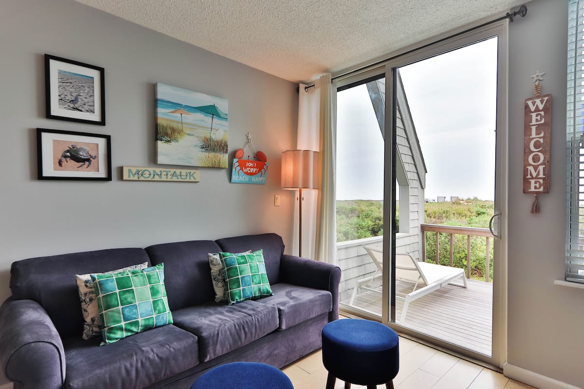 A room with a grey sofa, two green pillows, a framed picture of a beach, a 'Welcome' sign, and a view of a balcony with outdoor furniture.