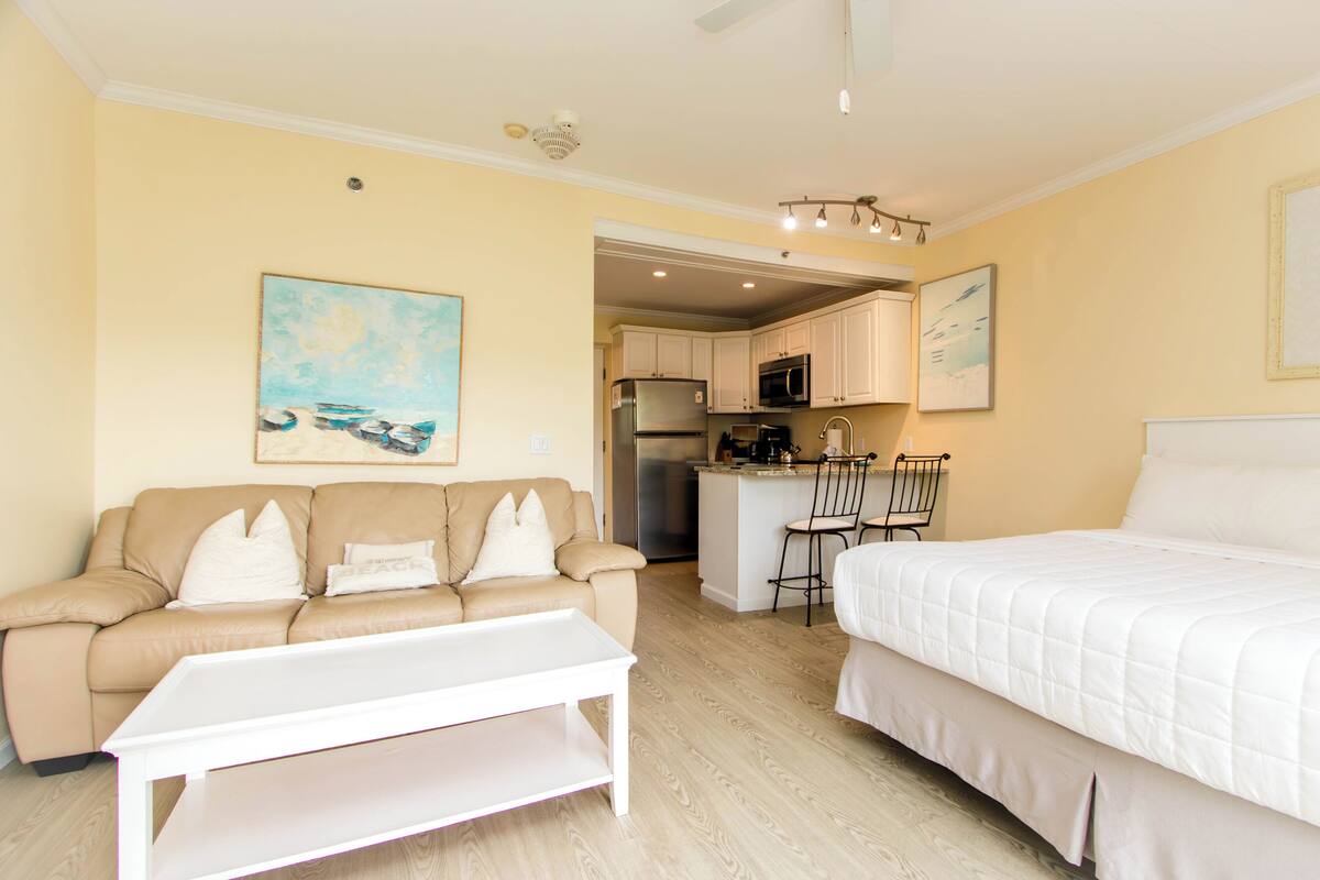 A hotel room with a bed, a sofa, a small table, a kitchenette, and a ceiling fan.
