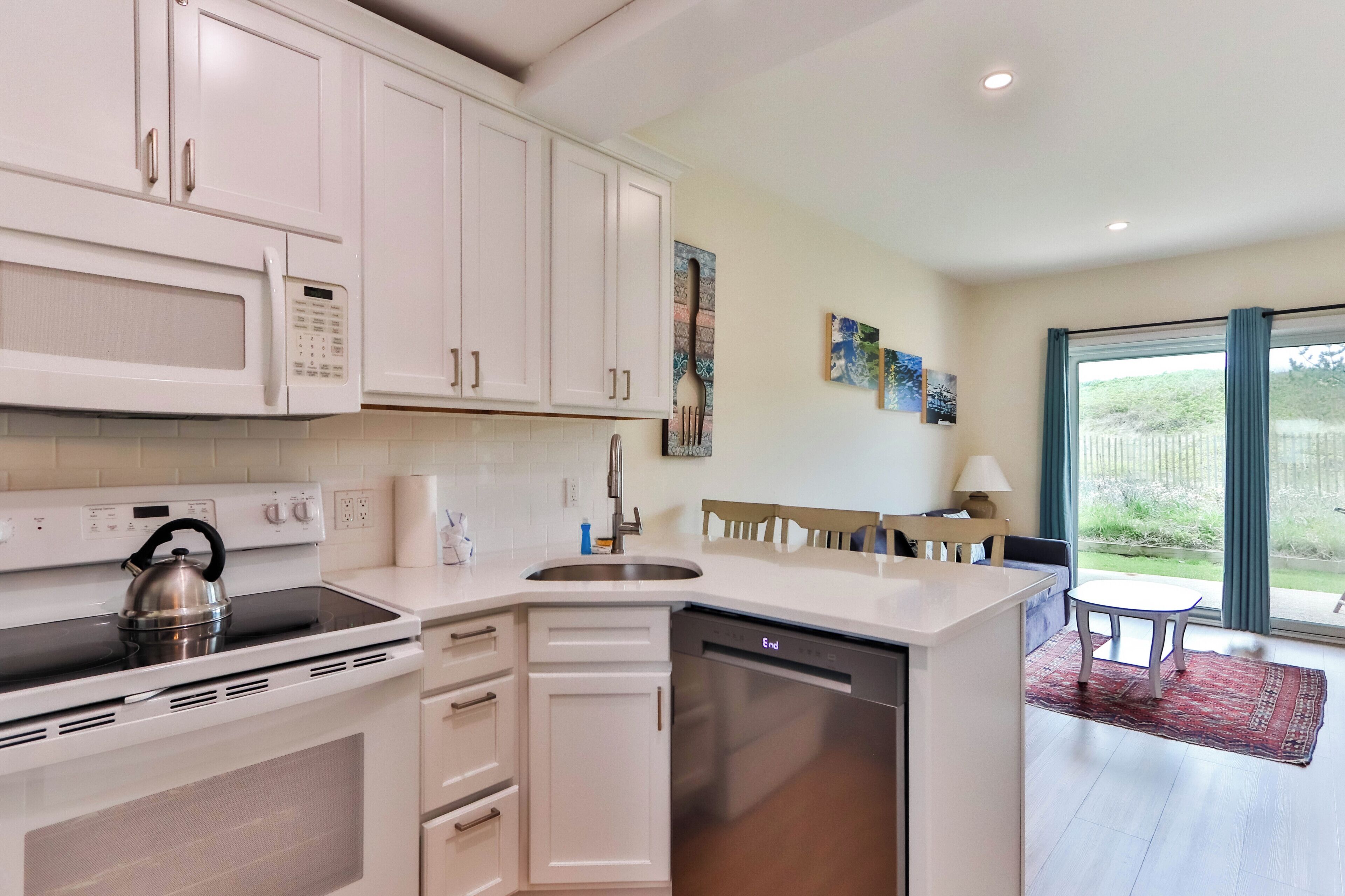 A modern kitchen with white cabinets, a stainless steel stove, and a dishwasher. There is a dining area with a table and chairs, and a window with a view of greenery.